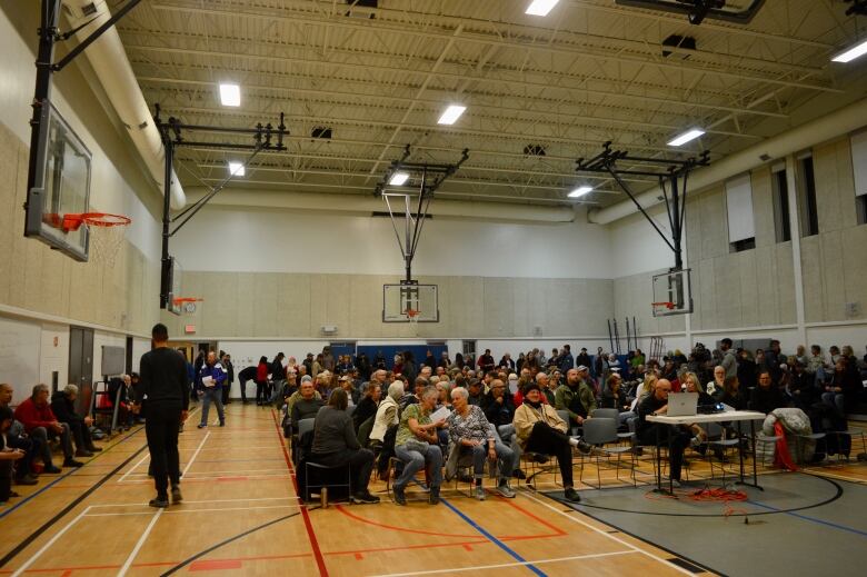 Many people sit in chairs that fill up a gymnasium. In the right of the frame is a table with a projector on it.