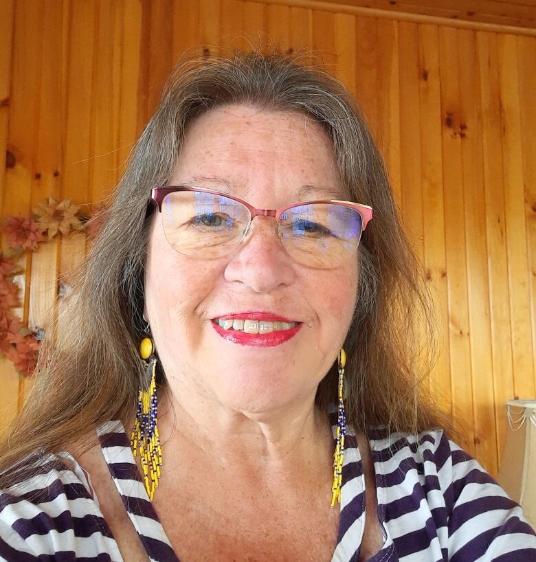 A Mi'kmaw woman with long brown and grey hair smiling, taking a selfie. She is wearing glasses, a striped shirt, and dangly yellow beaded earrings.