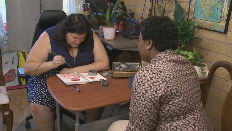 Woman leans down painting face on sketchpad as another woman sits across the table from her.