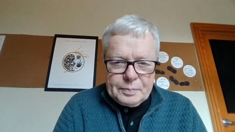 A man with white hear and glasses wears a green sweater. He is sitting in front of a wall.