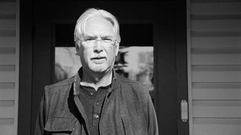 A black-and-white photo of a man outside a building. He has mid-length white hair, a goatee and glasses.