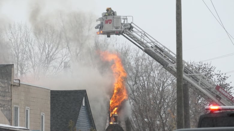 A large wave of flames come out of the top window of a 1 1/2-storey house. Firefighters spray water from above, in an aerial bucket.