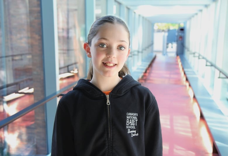 A girl in a dark hooded sweatshirt stands in a long hallway. 