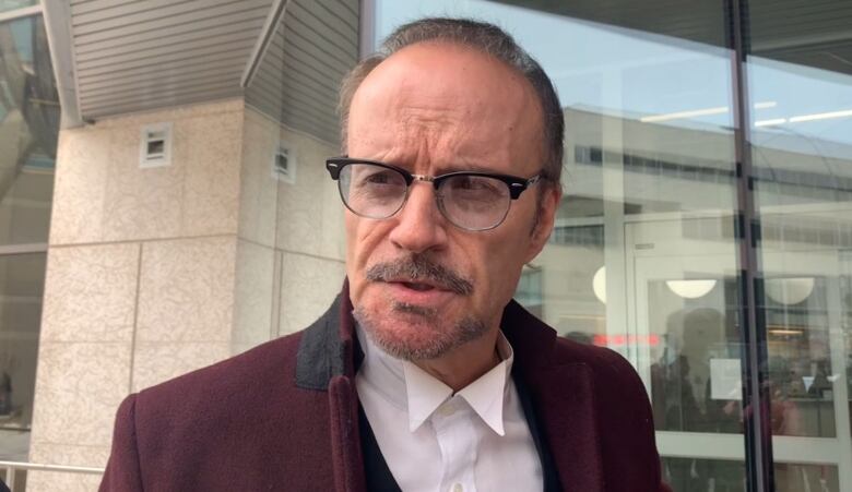 A man in a burgundy coat and glasses speaks outside a building.