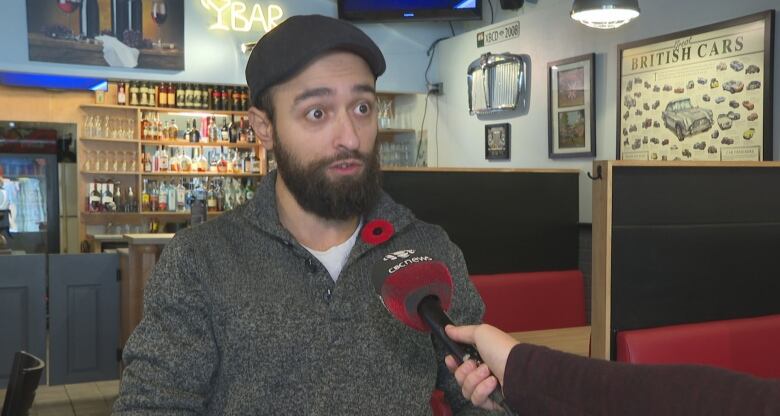 A bearded man wearing a British-style cloth cap and a grey sweater with a poppy stands in a British-themed room. 