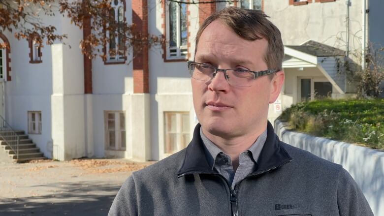 A white man with glasses and brown hair wearing a grey sweater stands outside with a large white building behind him