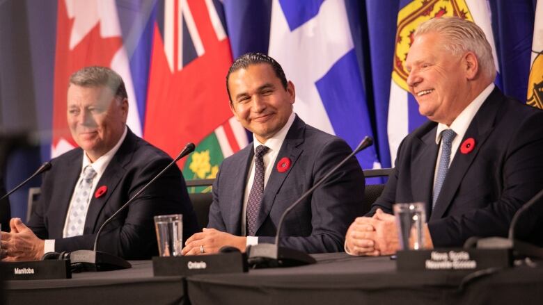 Three men in suits sit at a table, with provincial flags in the background.