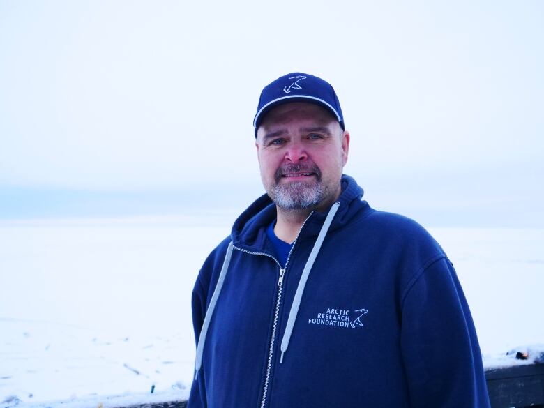 A man in a baseball cap and hoodie stands outside in Tuktoyaktuk, N.W.T., with snow in the background.