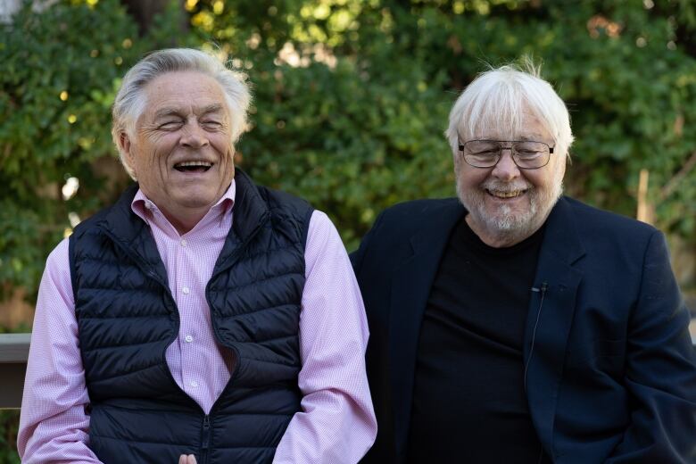 Two older, white-haired men laugh while sitting for a photograph.