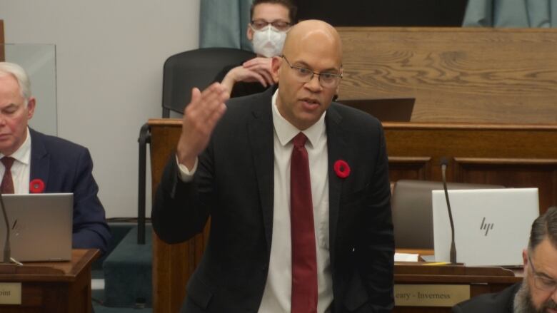 A man in a suit and tie stands and gestures toward the other side of the room, where other politicians are sitting.