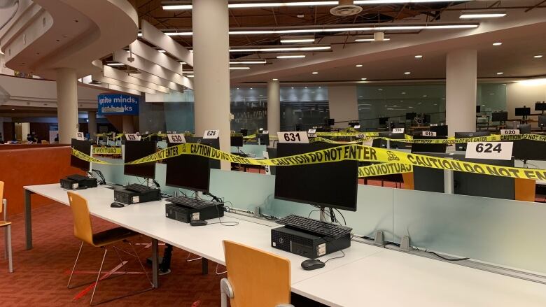 A row of desktop computers in an empty library are roped off by yellow tape.