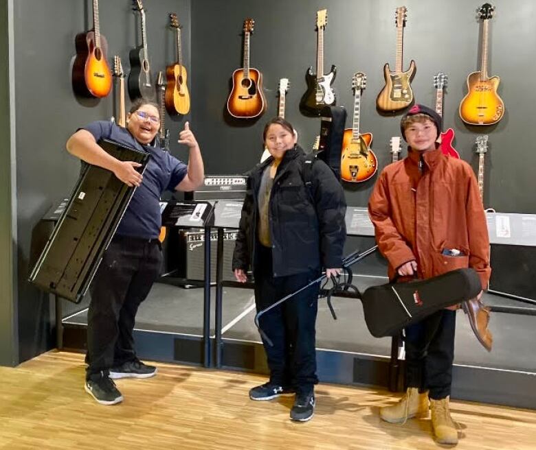 Three boys carrying their own instruments pose in front of a wall full of electric guitars.