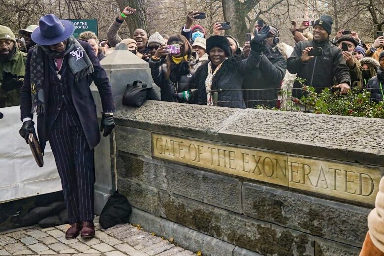 A man in a suit, hat and gloves stands next to a monument that says, 'Gate of the Exonerated.'