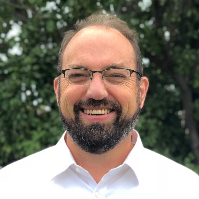 A man with glasses and a white collared shirt smiles at the camera