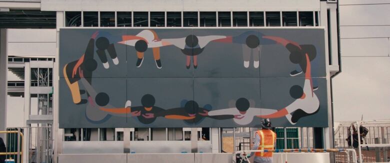 Still from Geoff McFetridge: Drawing a Life. View of the artist's mural (Us as a Measure of Openness) for the Westchester/Veterans Station in Inglewood, Calif. The artwork is a large panel installed on the exterior of a building. Minimalist in style, it depicts a circle of human figures, viewed from above. They are all joined arm in arm.