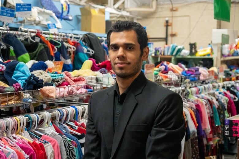 A man wearing a black suit jacket and dress shirt stands between racks of colourful clothing.