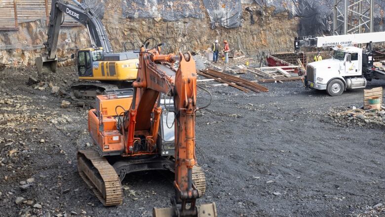 Orange and yellow piece of heavy construction equipment like front-end loaders sit in the bottom of a construction site with people in high-vis gear behind them