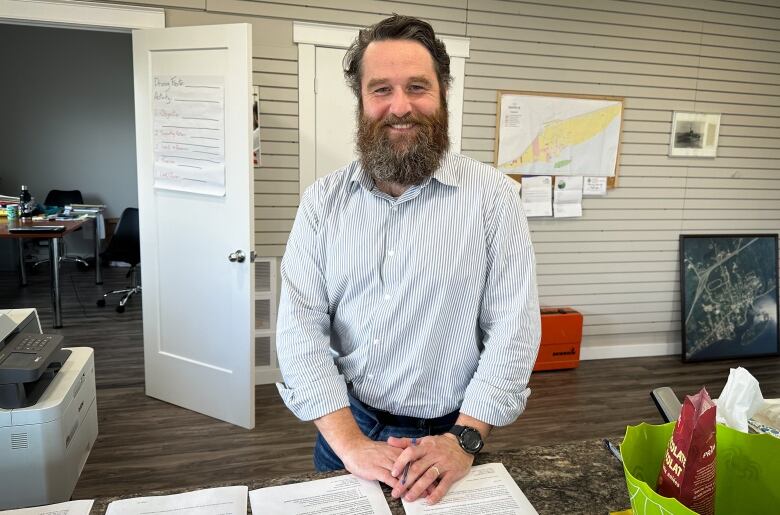 A man with brown hair and a bushy beard and white shirt stands behind a counter in an office.