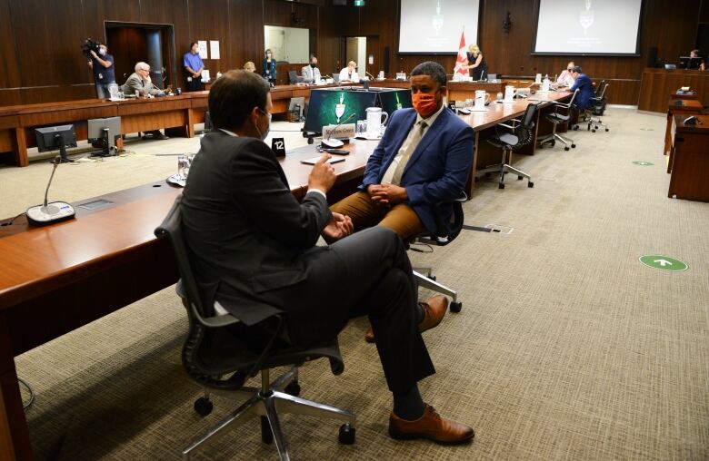 Two men in suits, one wearing an orange mask, sit at a board table and talk.