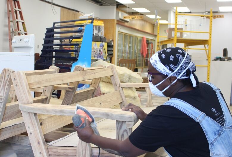 A woman wearing a respirator mask uses an electric sander on a bench, with other construction equipment in the background.