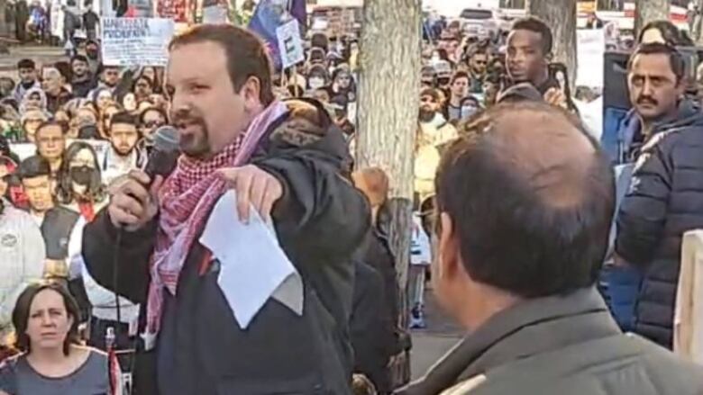 A man holding a microphone and a piece of paper addresses a crowd of protesters.