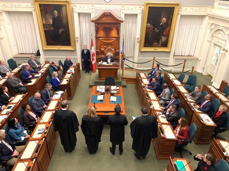 For people stand in front of a table with rows of people seated on either side.