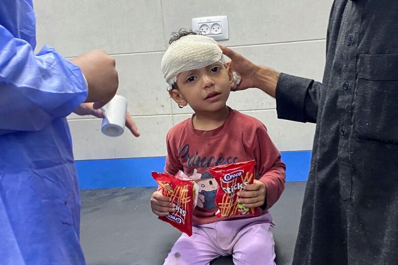 A small girl sits on a hospital table with a bandage wrapped around her forehead.