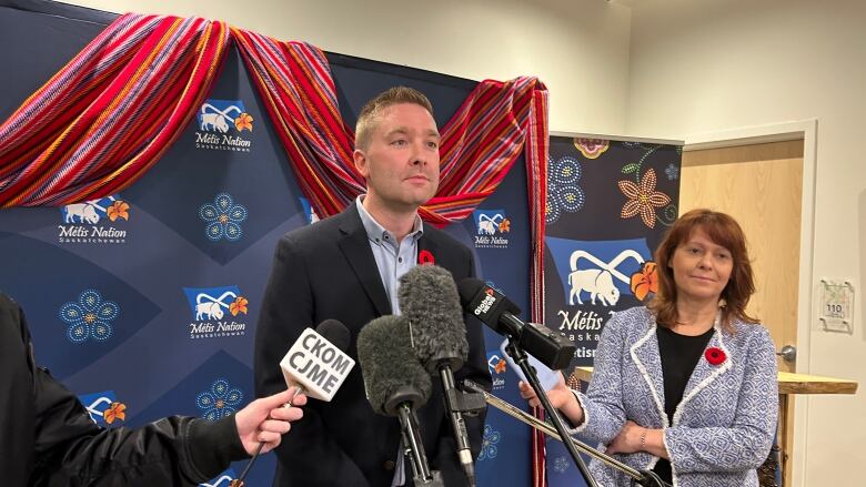 A man wearing a suit can be seen standing with a woman standing to his left. He is being interviews by reporters. 