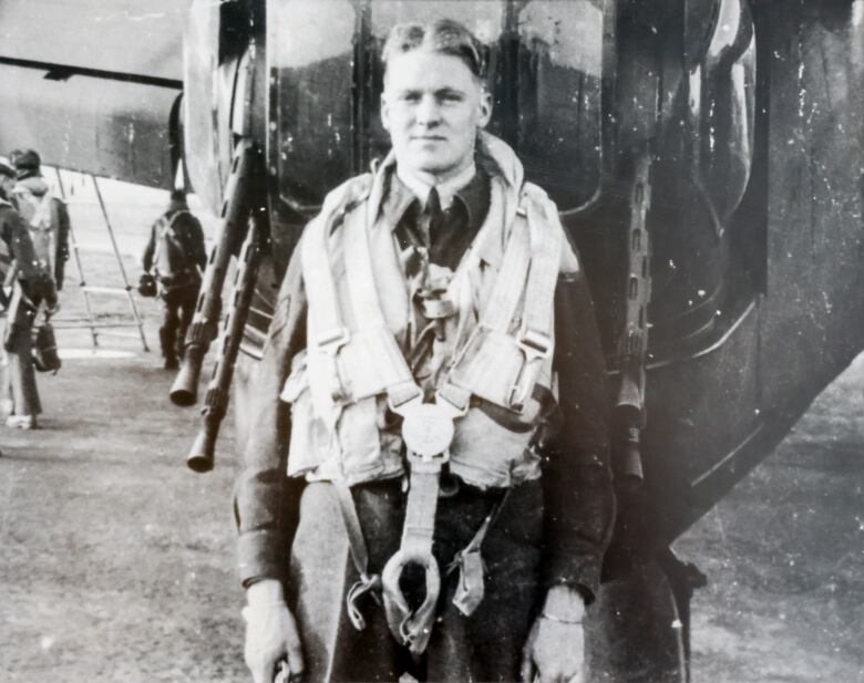 Black and white photo of a bomber crew airman from the Second World War. He is standing outside the plane, between the guns of tail turret.