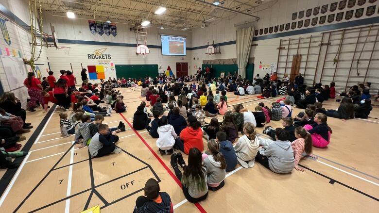 Several students sit in a school gymnasium, looking at a projection on a screen.