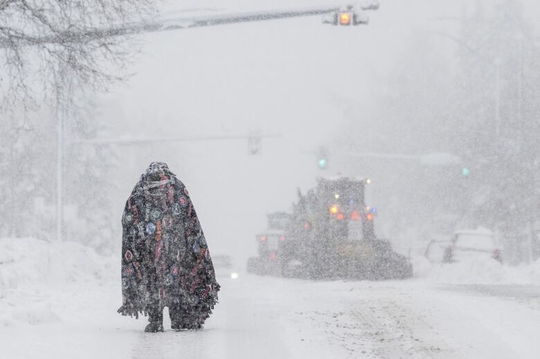 A person covered in a blanket walks down a street during a heavy snowstorm.