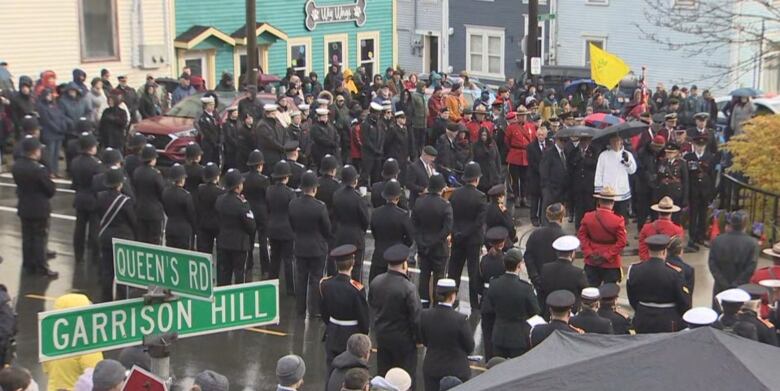 Military members along with police officers surround a man at a pulpit speaking. The event is outdoors and there appears to be rain. Large crowds of the public can be seen around the edges of the image, as well as a street sign showing the location. 