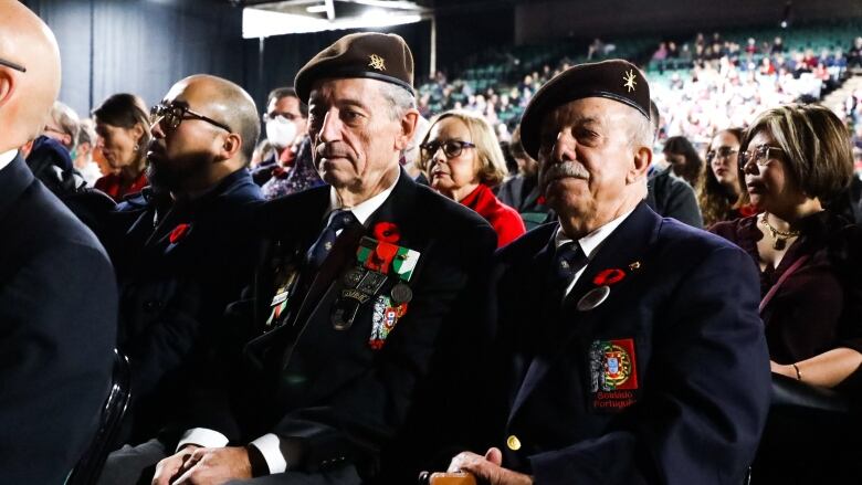 People wearing a uniform and poppies sit in a crowd.