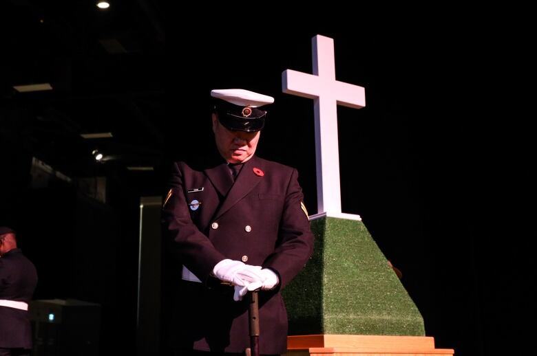 A person in a uniform wearing a poppy stands with his head bowed next to a white cross.