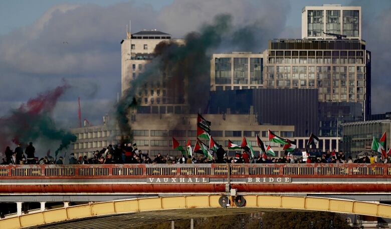 Protesters carrying flags and holding colourful smoke bombs walk across a bridge. 