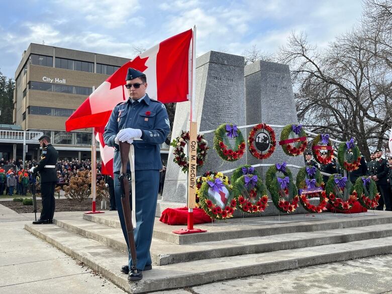 Two soldiers in blue uniforms hold rifles pointed downwards in front of a Canadian flag, with a stone war memorial behind them covered in wreaths and red poppies.