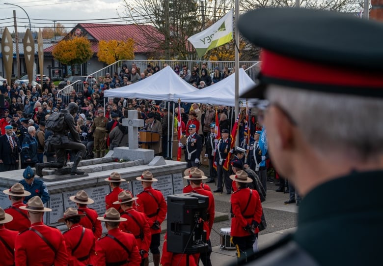 A number of  people wearing a red sash, along with members of the general public, gather around a cenotaph.