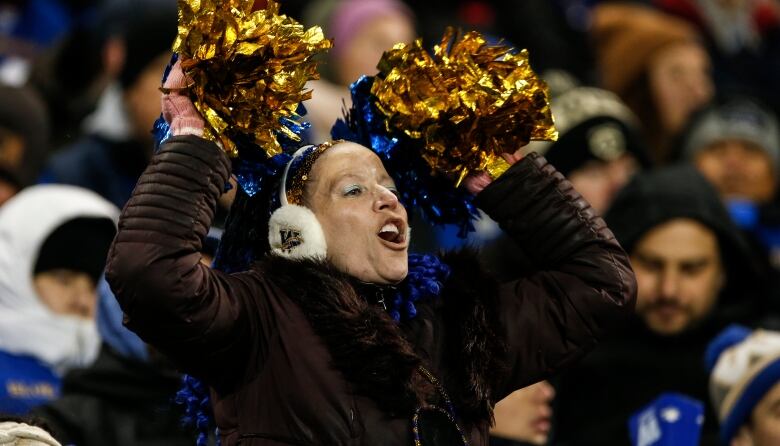 A woman standing in a crowd at a stadium cheers as she waves blue and gold pom-poms.