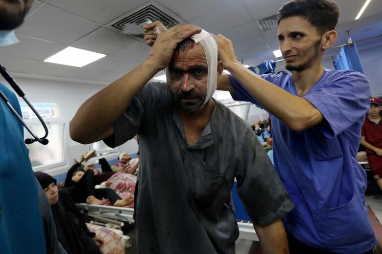 A person with blood on their face and a bandage around their head is treated by a person in scrubs inside a hospital.