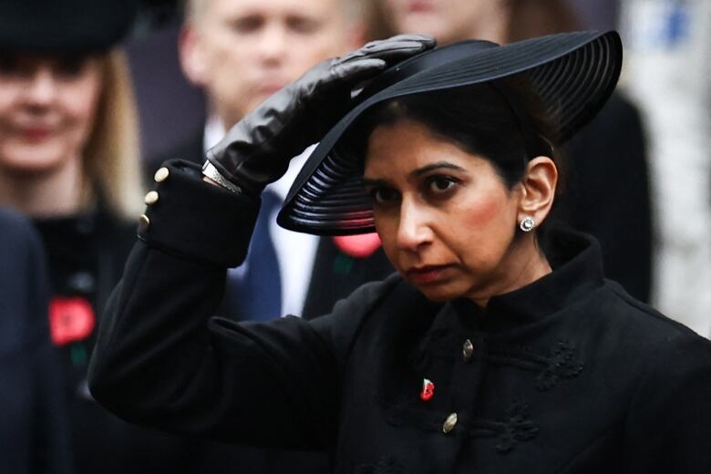 A woman in black holds a wide-brimmed hat to her head.