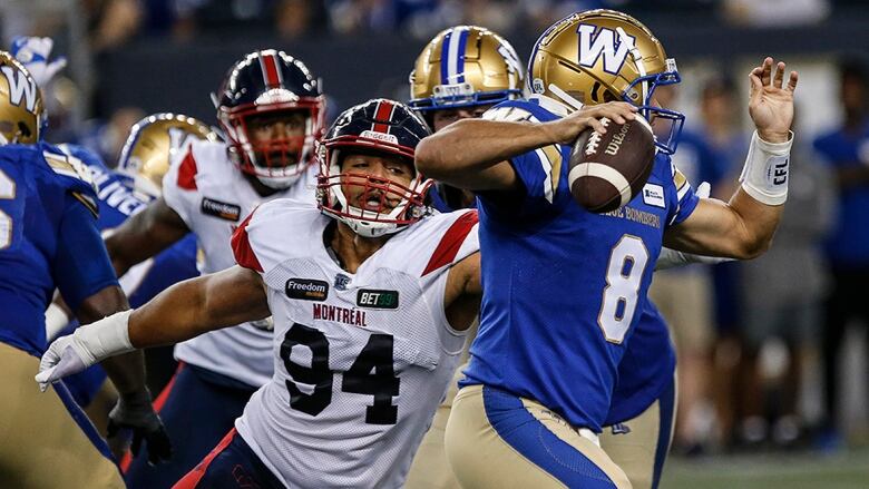 Men's quarterback holds football in right hand and is about to deliver a pass as an opposing player extends his left arm in an attempt to sack him in a CFL game.