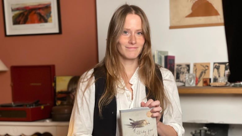 A woman with long auburn hair holds a book cover in her hands. She is standing in a living room with books in the background. 