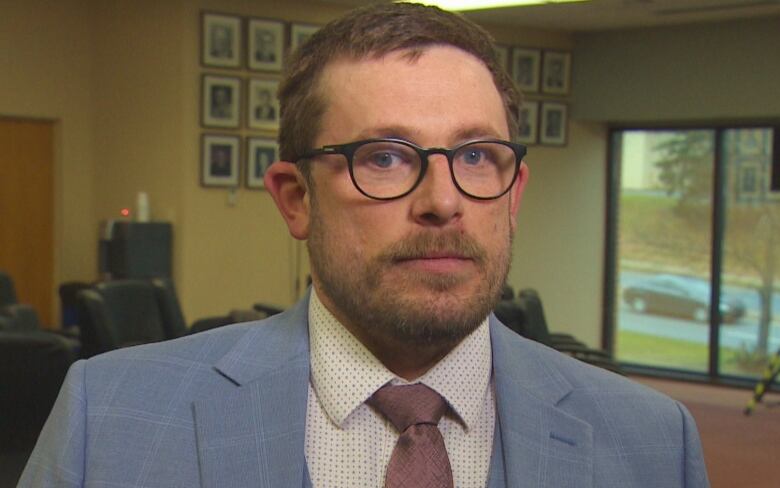 A man with short brown hair and black glasses. He is wearing a light blue suit jacket, a polka-dotted white dress shirt and a dusty pink tie.