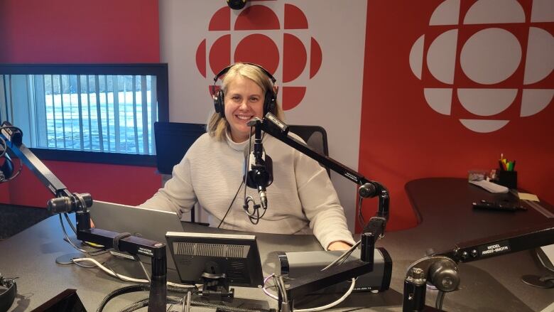 A blonde woman with a white sweater sits smiling behind a microphone inside a radio studio