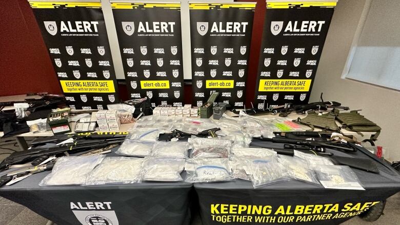 a table is pictured with many bags of white powder and  several rifles on it. 