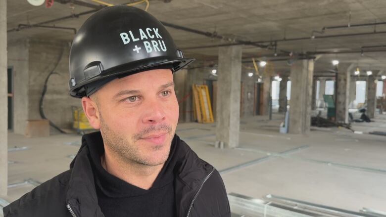 A man stands in a residential construction site, looking off-camera. He's wearing a black jacket and black hard hat.