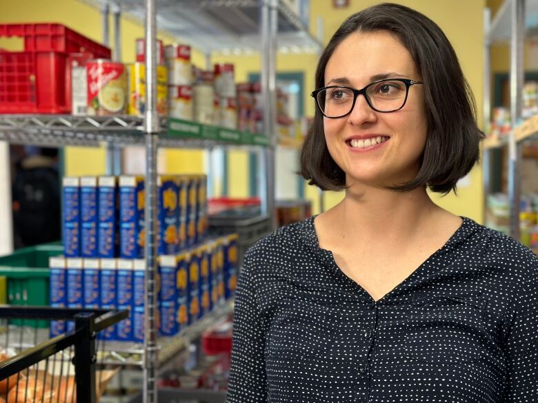 A woman with short, dark hair and glasses stands next to a shelf with food on it.