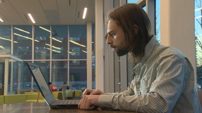 A man with a beard and shoulder-length hair looks at a laptop while sitting at a table indoors.