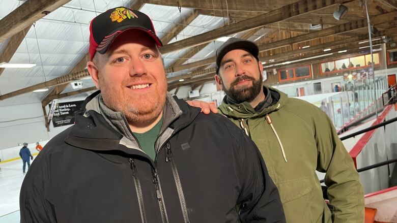 A man wearing a black jacket and a black baseball hat stands in front of another man in a green jacket and grey baseball hat. The man in the back has his right hand on the other man's left shoulder.