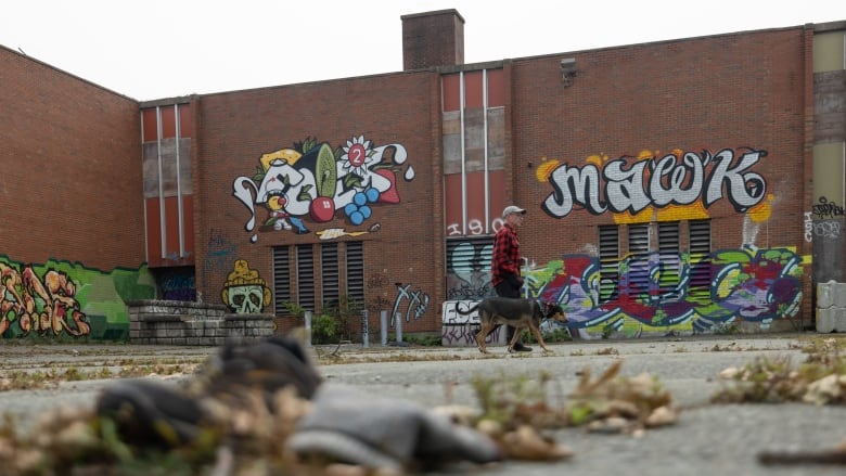 A man walks his dog past a brick building covered in graffiti.